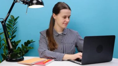 Kendine güvenen başarılı çalışan iş kadını 20 'li yaşlarda beyaz ofis masasında oturuyor dizüstü parmağını kendi üzerinde kullanıyor ve stüdyodaki mavi arkaplan duvarına izole edilmiş bir poz veriyor. İş konsepti