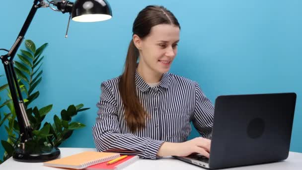 事业成功的女雇员20多岁快乐地坐在白写字台工作用笔记本电脑做电话手势说 拿回手指相机对着你 在工作室里摆出隔离的蓝色背景 — 图库视频影像