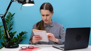 Hüsrana uğramış üzgün sekreter, sıradan gömlekli iş kadını. Beyaz ofis masasında bilgisayarlı kalem ve not defteriyle oturup mavi arka plan stüdyosunda izole bir şekilde poz veriyor.