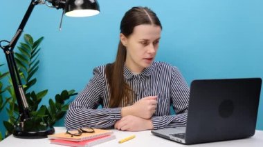 Başarılı çalışan iş kadını ofis masasında dizüstü bilgisayarla çalışır konferans görüşmesi yapar, gülümseyen genç kız kullanıcı el sallayarak internet görüntüsü çeker, mavi stüdyo arka planında izole olur.