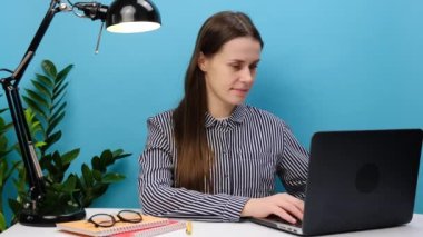 Başarılı çalışan 20 'li yaşlarda bir kadının portresi beyaz ofis masasında dizüstü bilgisayarla yoga yaparken el ele tutuşup rahatlamaya meditasyon yapmaya çalışarak stüdyonun mavi arka plan duvarında izole bir şekilde poz veriyor.
