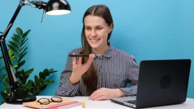 Sekreter çalışanı genç iş kadını portresi beyaz ofis masasında dizüstü bilgisayarla çalışıyor sesli mesajlar yolluyor ses konuşmalarıyla konuşuyor stüdyodaki mavi arkaplan duvarının üzerinde izole bir şekilde duruyor.