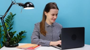 Gündelik gömlekli genç iş kadını ofisteki masa başı nokta dizüstü bilgisayarında internet kullanarak çalışıyor ve pastel mavisi stüdyo arka plan duvarında izole edilmiş şekilde poz veriyor. İş dünyası