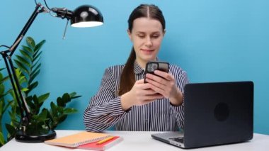 Güzel, başarılı bir çalışanın portresi beyaz ofis masasında cep bilgisayarlı, cep telefonlu, internette gezinen, pastel mavi arka plan duvarında izole bir şekilde poz veren genç bir kadın.