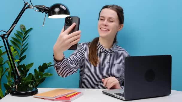 穿着休闲装的快乐秘书女员工的肖像 穿着便服坐在写字台工作 带着笔记本电脑在手机上自拍 在工作室的蓝色背景下显得孤立无援 业务概念 — 图库视频影像