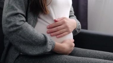 Close up of unknown pregnant woman sitting on comfy couch, puts palms on big belly feel unconditional love to unborn baby. Third trimester of pregnancy, happy maternity, expectant mother concept