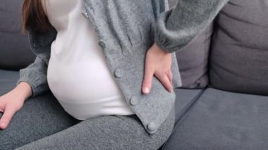 Close up side view of young pregnant woman massaging back muscles sitting on sofa, suffering from severe painful feelings, female 20s with third trimester big belly having joints strain in lower back