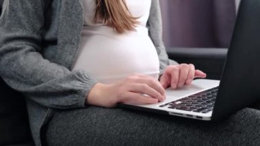 Close up of pregnant freelance employee woman sitting on cozy couch working from home, typing on laptop at big belly indoors, using online app, chatting, shopping on Internet, consulting doctor