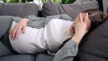 Unhappy tired young pregnant woman suffering from headache or negative thoughts, worrying about bad prenatal screening tests results, lying on couch at home. Feeling fear of childbirth concept