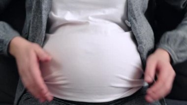 Close up of unrecognizable pregnant woman holding hands on belly and making heart shape sitting on couch. Lady feel love and care to her unborn baby enjoy beautiful period of being pregnant concept