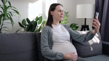 Smiling young pregnant woman sitting on couch holding cell phone in hands, distant web camera call using mobile application. Happy attractive female blogger recording video, sharing useful information