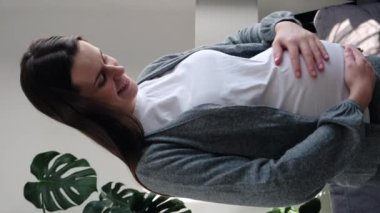 Vertical view smiling dreamy young beautiful mom posing in cozy indoors, holding hands on big belly, looking at window, enjoying calm sunny day, gift of life, happy pregnancy and maternity concept