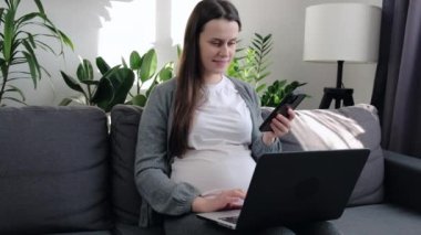 Happy beautiful young pregnant woman sitting on cozy couch using laptop and cellphone. Future mom expecting child relaxing at home in living room, dialing number and looking at smartphone screen