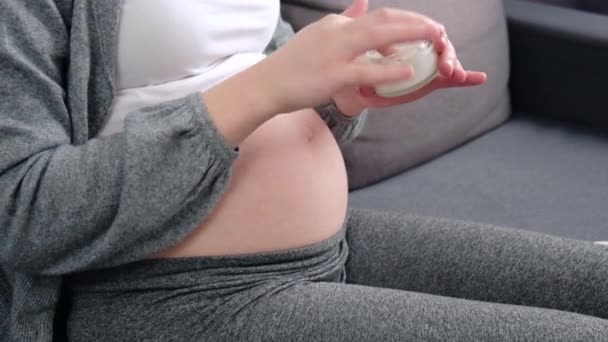 妊娠中の若い妊娠中の女性がストレッチマークを防止するために妊娠中の腹にスキンケアクリームを適用します 自宅で居心地の良いソファに座っている女性 妊産婦とケアの概念 — ストック動画