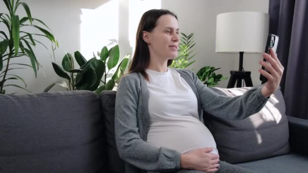 Χαμογελώντας Νεαρή Έγκυος Γυναίκα Κάθεται Στον Καναπέ Κρατώντας Κινητό Τηλέφωνο — Αρχείο Βίντεο