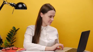 Portrait of cheerful woman in shirt holding small red heart sit work at desk with pc laptop, posing isolated over yellow color background wall in studio. Achievement business career lifestyle concept