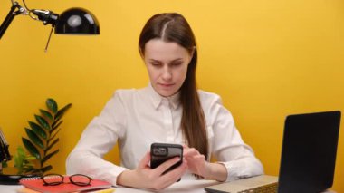 Portrait of happy successful employee business woman 20s sit work at office desk with laptop hold mobile cell phone do winner gesture clench fist, posing isolated over pastel yellow studio background