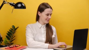 Smiling employee young woman 20s holding like counter template sitting at workplace with laptop, making post in social network and waiting for feedback, posing over yellow color background in studio