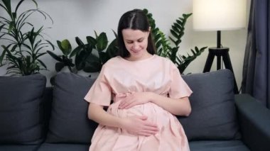 Portrait of smiling pretty young pregnant woman stroking big belly, happy looking at camera, sitting on cozy couch in living room, daydreaming at home. Hopeful candid positive lady enjoying pregnancy