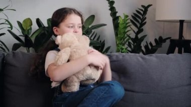Cute child hugging teddy bear and smiling, little girl playing with favorite toy sit on cozy sofa at home. Playful happy kid resting spending time alone embrace fluffy toy having. Childhood concept