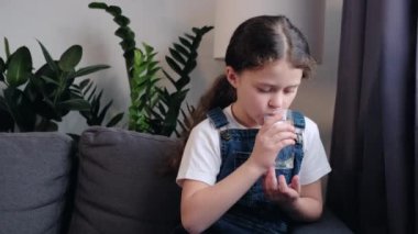 Gülümseyen küçük kız, cam gazsız temiz doğal mineral suyu içtiği için seviniyor. Küçük çocuk gülümseyerek yana bakıyor. Sağlıklı hissediyor. Sıcak kanepeye oturmuş susuzluğunu gideriyor.