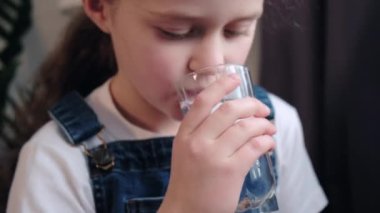 Close up of thirsty funny small cute kid girl drinking fresh pure water, refreshing during day or enjoying morning healthcare routine, sitting on cozy couch in living room at home. Selective focus