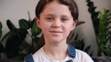 Close up portrait of adorable smiling little girl kid positive looking at camera, sitting alone on cozy couch at home, cute happy child with pretty face sit on sofa in living room. Concept childhood