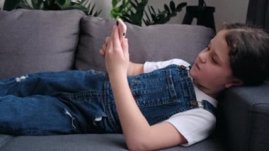 Addicted to modern tech adorable small kid girl lying on comfortable couch at home, involved in playing online mobile games alone. Joyful little school child having fun online in cozy living room