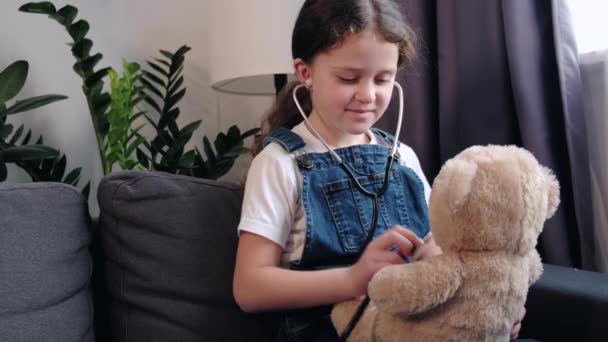 可爱的7岁小女孩拿着听诊器作为医生治疗坐在舒适的沙发上的充填玩具 笑着的小孩玩得开心医院听着毛绒绒的熊的心脏 梦想着未来的医学生涯 — 图库视频影像