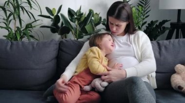 Close up of worried pregnant mother comforting solacing embrace crying little child daughter give care and protection sitting on sofa. Loving concerned mom hug sad small girl consoling kid concept