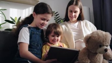 Close up of pretty two little daughters reading book with beautiful young pregnant mom, sitting on cozy sofa in living room together, enjoying spending time at home. Childhood and motherhood concept