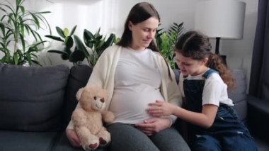 Motherhood concept. Expecting young mom and happy pretty little daughter hugging, relaxing on sofa together, touching pregnant mothers belly, talking, discussing planned younger sibling, baby birth