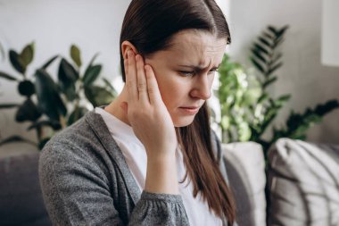 Close up portrait of upset young girl holding painful ear, suddenly feeling strong ache. Unhealthy caucasian woman 20s suffering from painful otitis sitting on sofa at home. Health problems concept clipart