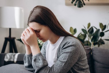 Thoughtful young female sit on couch lean forward on folded hands try to keep emotions under control. Sad woman think of overcoming bad life event ponder on hard decision. Critical situation concept