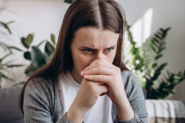Close up of stressful sad young caucasian woman sitting alone on couch in living room covering face with hands. Emotional brunette female 20s feeling upset and unhappy at home. Depression concept