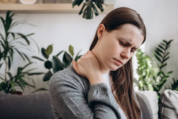 Selective focus of unhealthy young woman having neck pain sitting on couch at home. Upset girl has pain from muscles. Cervical osteochondrosis, tired overworked female, sedentary lifestyle concept