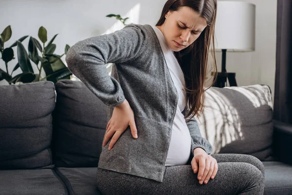 Side view of upset pregnant young female suffering lower back pain sitting on sofa in living room. Sad future mother suffer from spine muscle ache backache. Concept pregnancy, motherhood, health