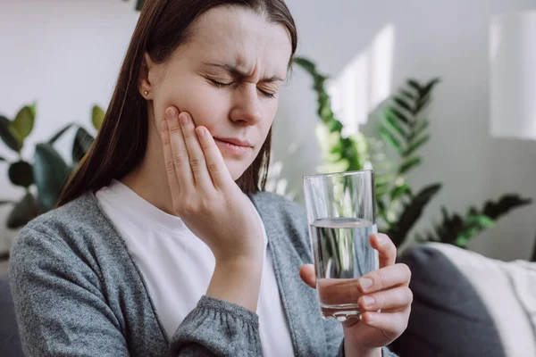 Portrait of young woman feel terrible toothache after drink cold water. Female sitting on couch at home touching cheek, feel hurt and suffering from sensitive tooth ache. Pain and cavities concept