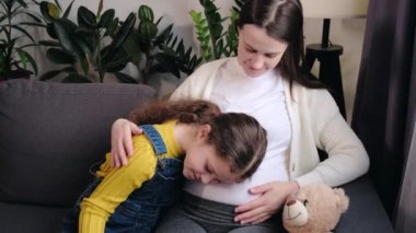 Close up of pretty little daughter hugging and listening pregnant mother belly, bonding together at home. Young happy pregnant mom embracing kid girl sitting on cozy sofa. Pregnant mother and child