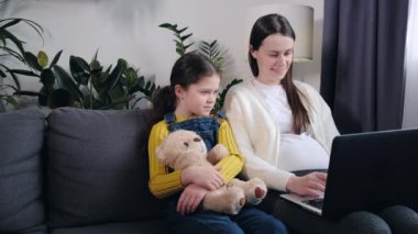 Cute young pregnant mother with pretty little daughter using laptop at home in living room. Smiling mom watching cartoons on computer with small girl kid sitting on comfy couch. Motherhood concept