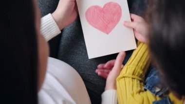 Top view of mother hugging cute little daughter, thanking for gift, sitting on sofa at home, small girl child congratulating mum with birthday or mothers day, presenting greeting card with red heart