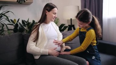 Happy young mom and pretty little daughter child hugging, relaxing on couch together, touching pregnant mother's belly, talking, discussing planned younger sibling, baby birth. Motherhood concept