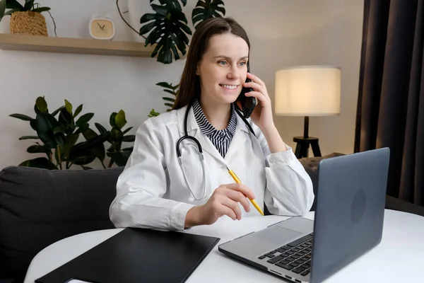 Smiling cute young female doctor physician holding cell phone talking on mobile, using laptop. Healthcare professional answering call giving remote consultation on smartphone making appointment notes