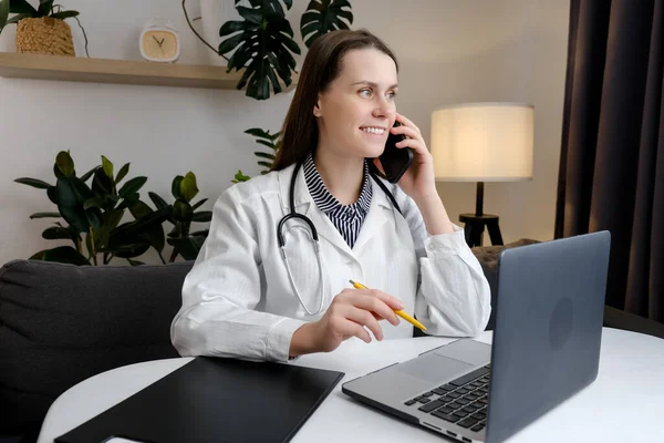 Smiling young woman physician talk by phone before pc. Happy female doctor in coat consulting person by phone fill in files on laptop describe complaints from patient words give advice prescriptions