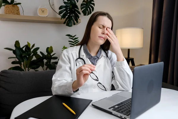 Upset young doctor female wearing uniform and stethoscope therapist physician sitting at table suffering from eye strain, dry eye syndrome, massaging eyelids, headache. Stress and frustration concept