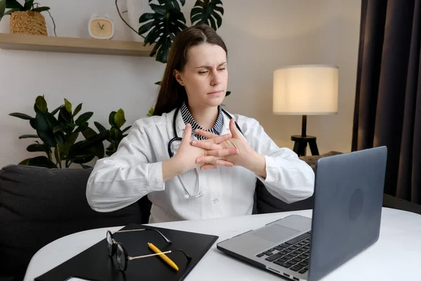 Upset doctor at computer experiences pain and discomfort in hands. Concentrated female general practitioner working at laptop typing on keyboard takes break and kneading fingers. Arthritis concept
