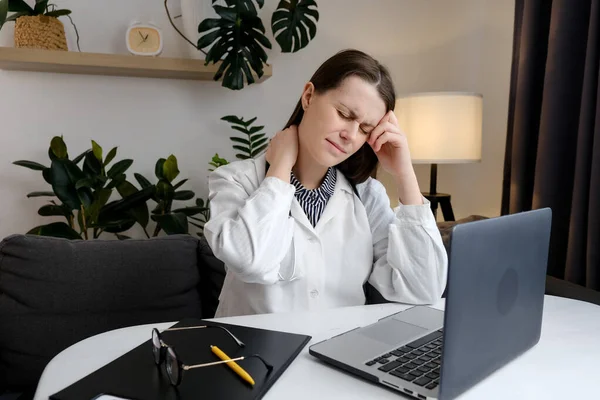 Tired sad female doctor at computer experiences pain and discomfort in neck. Concentrated woman general practitioner in office working at laptop typing on keyboard takes break and touching shoulder