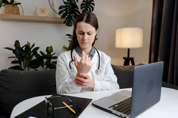 Tired doctor at computer experiences pain and discomfort in hands. Concentrated woman general practitioner working at laptop and suffering from wrist pain sitting at table in clinic. Arthritis concept