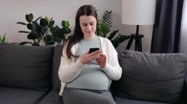 Modern akıllı telefonlu mutlu hamile kadın oturma odasında rahat bir kanepede dinlenirken sosyal medyayı kontrol ediyor. Neşeli, çekici bir kadın. Evde parmaklarıyla kaydırıyor ve dokunuyor.