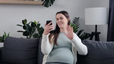 Çekici genç hamile kadın rahat bir koltukta oturuyor cep telefonunu elinde tutuyor, mobil uygulama kullanarak uzak bir web kamerasından arıyor. Müstakbel mutlu anne videosu, faydalı bilgileri paylaşıyor.